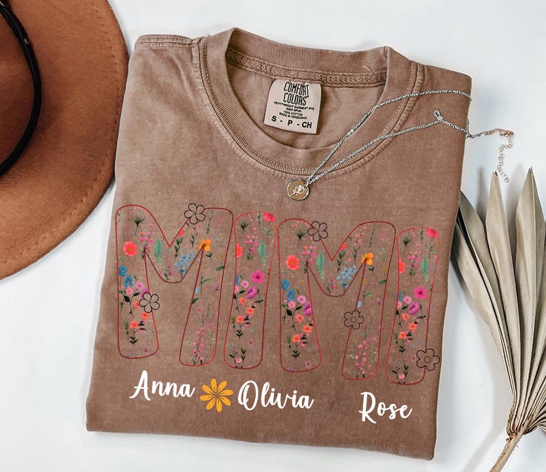Pode incluir: Camiseta marrom com a palavra "MAMA" em um design floral. Os nomes "Anna", "Olivia" e "Rose" s&atilde;o impressos abaixo. Um colar de prata com um pingente est&aacute; na camiseta.