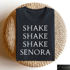May include: Black t-shirt with the words "SHAKE SHAKE SHAKE SENORA" printed in white. The shirt is displayed on a round, woven, light brown surface. The shirt is a crew neck style.