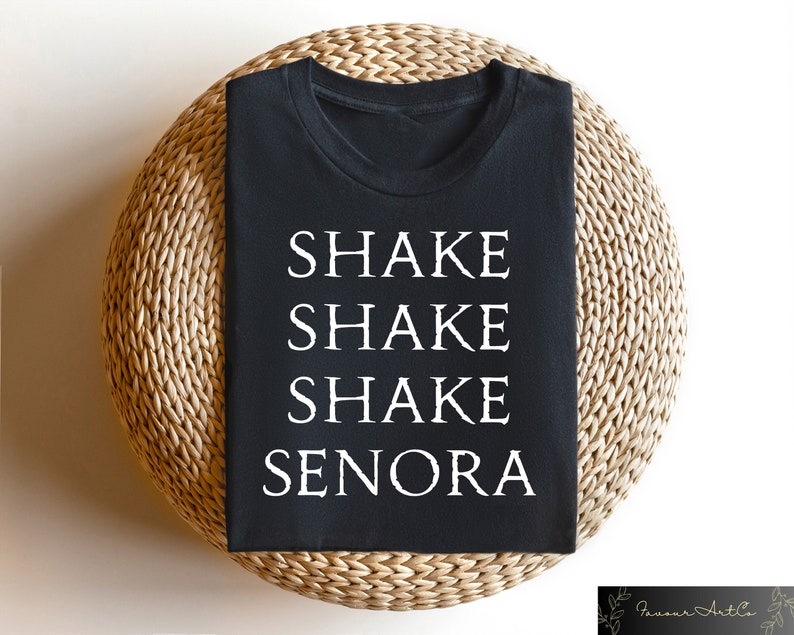 May include: Black t-shirt with the words "SHAKE SHAKE SHAKE SENORA" printed in white. The shirt is displayed on a round, woven, light brown surface. The shirt is a crew neck style.