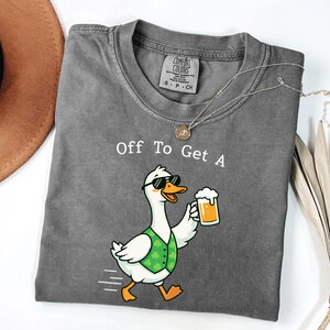 May include: A gray t-shirt with the text "Off To Get A" above a cartoon goose wearing sunglasses and a green vest, holding a beer. The goose is walking with motion lines.
