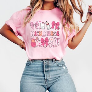 Peut inclure: T-shirt rose avec le texte "NICU NURSE" en lettres roses et blanches. Le t-shirt pr&eacute;sente une vari&eacute;t&eacute; d'illustrations roses et blanches d'articles pour b&eacute;b&eacute;s, notamment un biberon, une sucette, un st&eacute;thoscope et un c&oelig;ur.