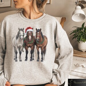 May include: A gray sweatshirt with a Christmas-themed design featuring three horses wearing Santa hats and holly wreaths. The horses are standing side-by-side, with the white horse on the left, the brown horse in the middle, and the black horse on the right.