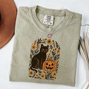 May include: A sage green t-shirt featuring a black cat and jack-o'-lantern design. The cat sits beside the pumpkin, surrounded by floral elements. A silver necklace with a pendant rests on the shirt. The shirt has a Comfort Colors tag.