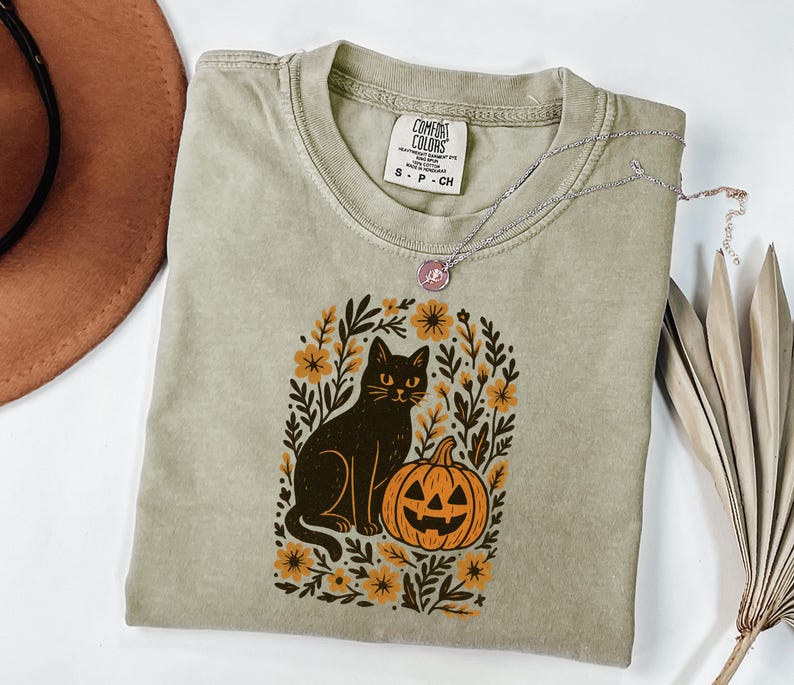 May include: A sage green t-shirt featuring a black cat and jack-o'-lantern design. The cat sits beside the pumpkin, surrounded by floral elements. A silver necklace with a pendant rests on the shirt. The shirt has a Comfort Colors tag.