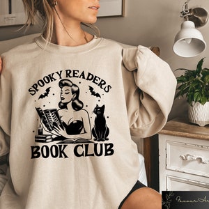 May include: A beige sweatshirt with a black graphic design featuring a woman reading a book with a black cat beside her. The text on the graphic reads "Spooky Readers Book Club".