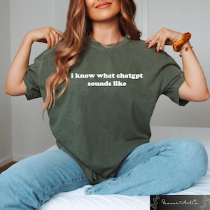 May include: An olive green t-shirt with the text "i know what chatgpt sounds like" in white. The shirt is worn with light blue jeans. The style is casual and comfortable.