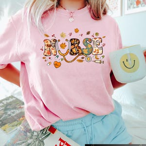 Pode incluir: Camiseta rosa com um gr&aacute;fico colorido de "ENFERMEIRA" que apresenta folhas de outono e s&iacute;mbolos m&eacute;dicos. O gr&aacute;fico inclui um estetoscopio, p&iacute;lulas, um cora&ccedil;&atilde;o e uma seringa.