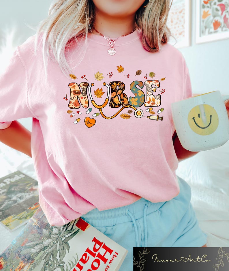 Pode incluir: Camiseta rosa com um gr&aacute;fico colorido de "ENFERMEIRA" que apresenta folhas de outono e s&iacute;mbolos m&eacute;dicos. O gr&aacute;fico inclui um estetoscopio, p&iacute;lulas, um cora&ccedil;&atilde;o e uma seringa.