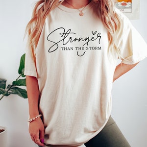 May include: A white t-shirt with the text "Stronger than the storm" in black cursive lettering with a small heart symbol.