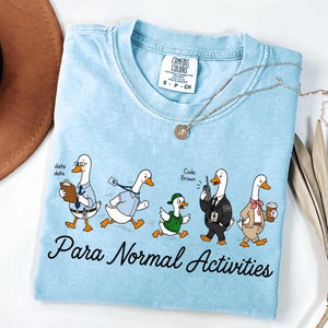 May include: A light blue t-shirt featuring a graphic of five ducks dressed in various outfits with the text "Para Normal Activities" printed below. The ducks are depicted as an office worker, a runner, a person in a hoodie, security, and a professional.