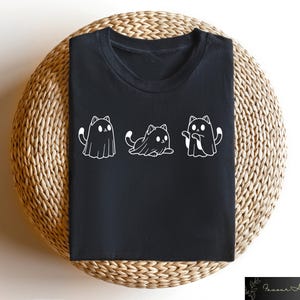 Pu&ograve; includere: Maglietta nera con una grafica bianca di tre gatti vestiti da fantasmi. I gatti sono in fila, con il gatto al centro seduto e i gatti ai lati in piedi.