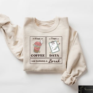 May include: Beige sweatshirt with a graphic design of a coffee cup and a checklist. The text reads "~First~ Coffee ~Then~ Data I AM EARNING A: Break"