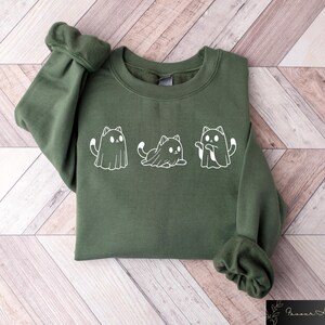 Pu&ograve; includere: Una felpa verde con una grafica bianca di tre gatti vestiti da fantasmi. I gatti indossano costumi da fantasma con occhi e bocche.