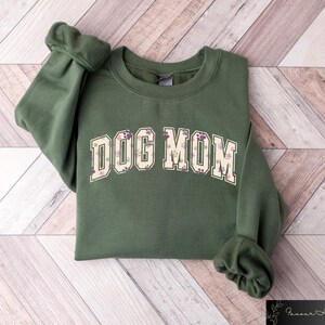May include: A green sweatshirt with the text "DOG MOM" in a floral print. The text is in a large, bold font and is centered on the sweatshirt.