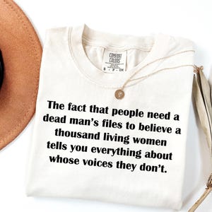 May include: Cream-colored t-shirt with the text "The fact that people need a dead man's files to believe a thousand living women tells you everything about whose voices they don't." A gold necklace is draped on the shirt.