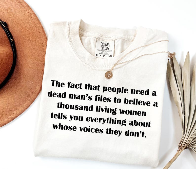 May include: Cream-colored t-shirt with the text "The fact that people need a dead man's files to believe a thousand living women tells you everything about whose voices they don't." A gold necklace is draped on the shirt.