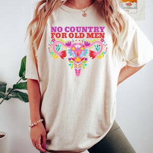 May include: A white t-shirt with the text "NO COUNTRY FOR OLD MEN" in pink and purple lettering. A colorful floral design of a uterus is centered on the shirt.