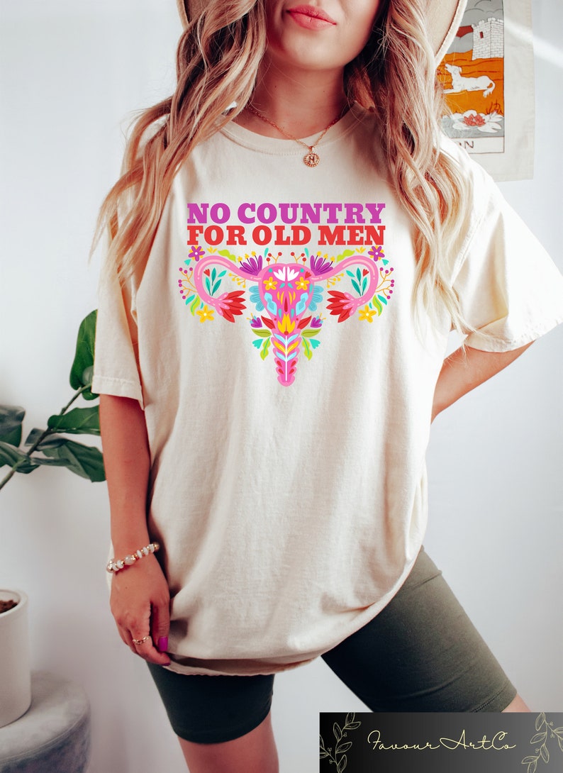 May include: A white t-shirt with the text "NO COUNTRY FOR OLD MEN" in pink and purple lettering. A colorful floral design of a uterus is centered on the shirt.