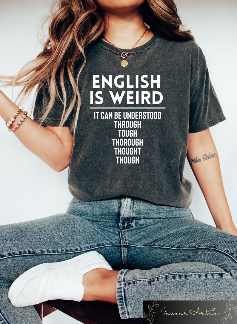 May include: A dark gray t-shirt with the text "ENGLISH IS WEIRD" and the words "IT CAN BE UNDERSTOOD THROUGH TOUGH THOROUGH THOUGHT THOUGH" printed in white.
