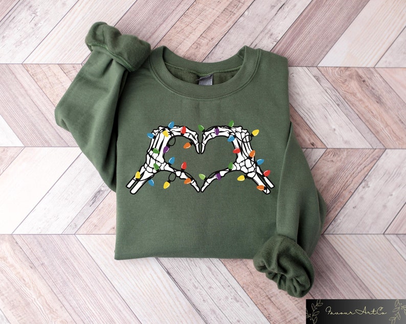 May include: An olive green sweatshirt featuring a heart made of white skeleton hands, adorned with colorful string lights. The design is centered on the chest. The sleeves are cuffed. The sweatshirt is displayed on a light wood surface.