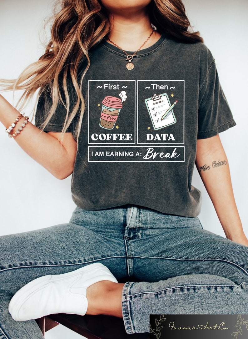 May include: A gray t-shirt with a graphic design of a coffee cup and a checklist. The text on the shirt reads "First Coffee, Then Data, I am earning a: Break".