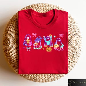 May include: Red t-shirt with a graphic of five neon ghosts holding pumpkins and candy. The ghosts are wearing hats and scarves. The graphic is surrounded by bows and stars.