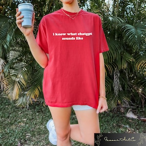 May include: A red t-shirt with the white text "i know what chatgpt sounds like". The shirt has a loose fit. A person is holding a light blue collapsible cup. The background features green foliage.