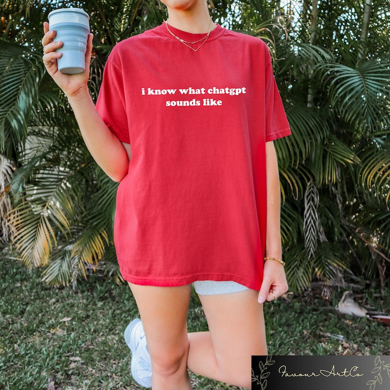 May include: A red t-shirt with the white text "i know what chatgpt sounds like". The shirt has a loose fit. A person is holding a light blue collapsible cup. The background features green foliage.