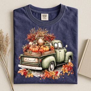 May include: A navy blue t-shirt featuring a graphic of a light green vintage pickup truck carrying a wooden crate filled with orange pumpkins, wheat, and red berries. The truck tailgate displays the text Autumn vibes, surrounded by fallen autumn leaves.