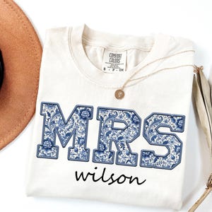 Puede incluir: Camiseta color crema con la palabra "MRS" en letras florales azules grandes y el nombre "wilson" en cursiva. Un collar de oro con un colgante descansa sobre la camiseta. La camiseta es de la marca Comfort Colors.