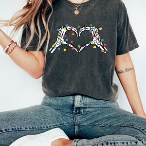 May include: Dark gray t-shirt featuring a heart made of white skeleton hands, adorned with colorful Christmas lights. The shirt is paired with blue jeans and white sneakers. The overall theme is a blend of Halloween and holiday.