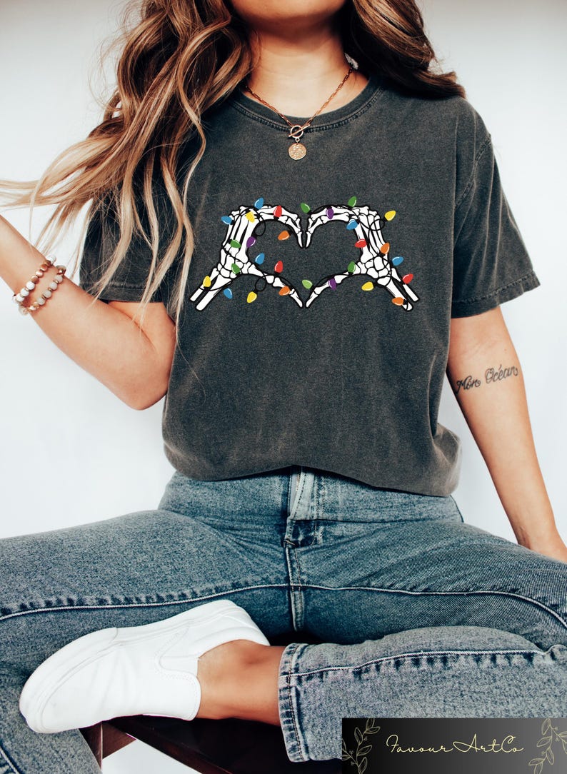 May include: Dark gray t-shirt featuring a heart made of white skeleton hands, adorned with colorful Christmas lights. The shirt is paired with blue jeans and white sneakers. The overall theme is a blend of Halloween and holiday.