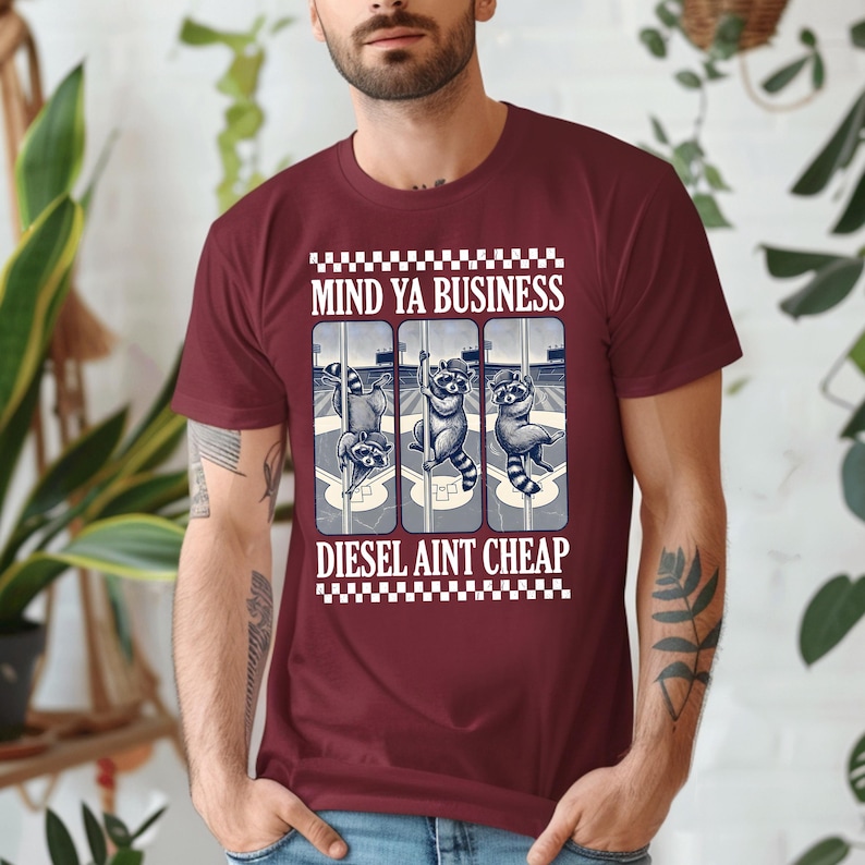 May include: Maroon t-shirt with the text "MIND YA BUSINESS" above a graphic of three raccoons in a baseball-themed design. Below the graphic is the text "DIESEL AINT CHEAP". The shirt has a crew neck and short sleeves.
