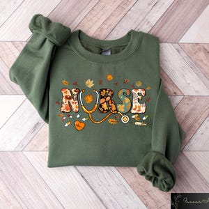Pode incluir: Uma camisa de moletom verde com a palavra "NURSE" escrita em letras coloridas com tema de outono. As letras s&atilde;o decoradas com folhas, flores e um estetosc&oacute;pio.