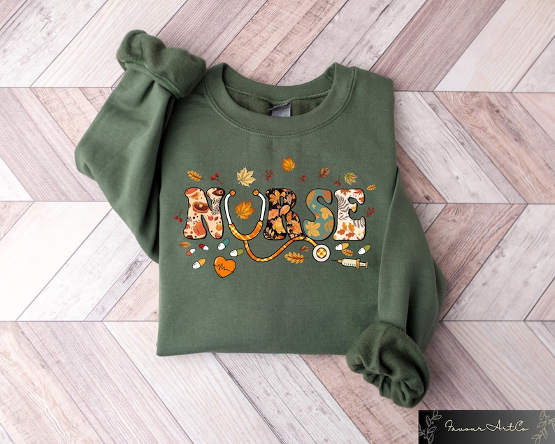 Pode incluir: Uma camisa de moletom verde com a palavra "NURSE" escrita em letras coloridas com tema de outono. As letras s&atilde;o decoradas com folhas, flores e um estetosc&oacute;pio.