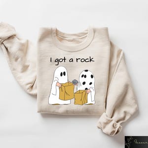 May include: A tan sweatshirt with the text "I got a rock" and a cartoon image of two ghosts trick-or-treating. One ghost is holding a rock in a brown paper bag.