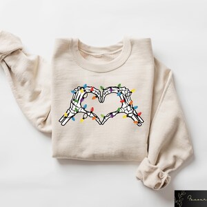 May include: Beige sweatshirt featuring a heart made of skeleton hands with colorful Christmas lights. The graphic is centered on the front of the long-sleeved, crew neck top. The sleeves are rolled up.