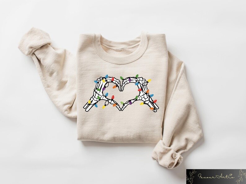 May include: Beige sweatshirt featuring a heart made of skeleton hands with colorful Christmas lights. The graphic is centered on the front of the long-sleeved, crew neck top. The sleeves are rolled up.