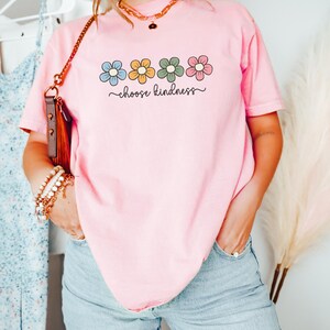 Puede incluir: Camiseta rosa claro con un dise&ntilde;o floral en azul, amarillo, verde y rosa, y la frase "choose kindness" en letra cursiva. La camiseta se combina con unos vaqueros azul claro.