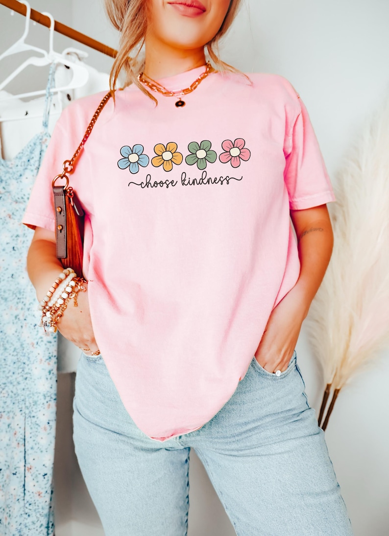 Puede incluir: Camiseta rosa claro con un dise&ntilde;o floral en azul, amarillo, verde y rosa, y la frase "choose kindness" en letra cursiva. La camiseta se combina con unos vaqueros azul claro.