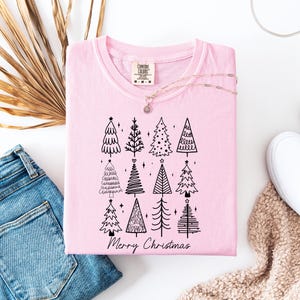 May include: A pale pink t-shirt featuring a black illustration of Christmas trees and the text "Merry Christmas." The shirt is displayed with blue jeans, white trainers, and a gold necklace.