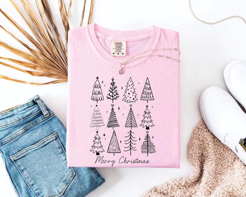 May include: A pale pink t-shirt featuring a black illustration of Christmas trees and the text "Merry Christmas." The shirt is displayed with blue jeans, white trainers, and a gold necklace.