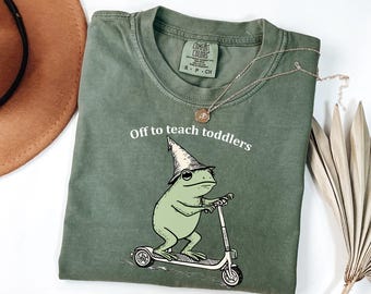 Könnte beinhalten: Olivgrünes T-Shirt mit dem Text "Off to teach toddlers" und einem Cartoon-Frosch mit Zaubererhut, der einen Roller fährt. Eine goldene Halskette und ein brauner Hut sind ebenfalls zu sehen.