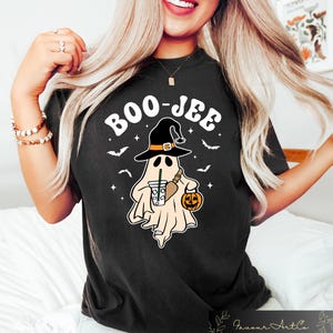 Może przedstawiać: Czarny T-shirt z białym nadrukiem przedstawiającym ducha w czarodziejskim kapeluszu trzymającego dynię i kubek kawy. Tekst "BOO-JEE" jest napisany na biało nad duchem.