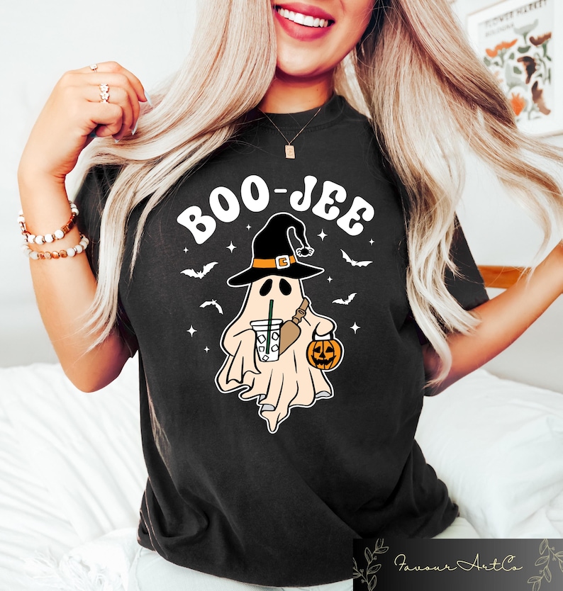 Może przedstawiać: Czarny T-shirt z białym nadrukiem przedstawiającym ducha w czarodziejskim kapeluszu trzymającego dynię i kubek kawy. Tekst "BOO-JEE" jest napisany na biało nad duchem.