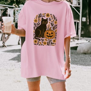 May include: A light pink t-shirt featuring a black cat and jack-o'-lantern graphic. The cat sits beside the pumpkin, surrounded by yellow flowers and green leaves. The shirt is a loose fit.