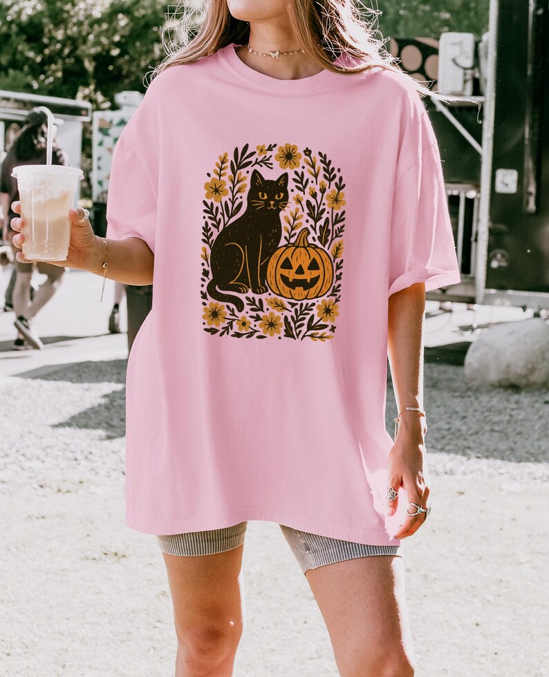 May include: A light pink t-shirt featuring a black cat and jack-o'-lantern graphic. The cat sits beside the pumpkin, surrounded by yellow flowers and green leaves. The shirt is a loose fit.