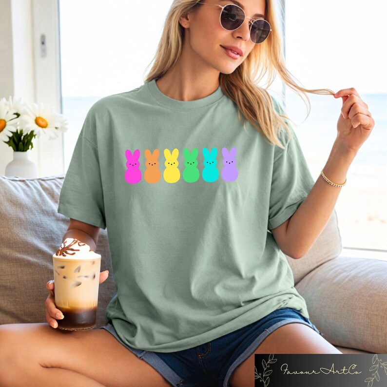 K&ouml;nnte beinhalten: Ein hellgr&uuml;nes T-Shirt mit einer Reihe von sechs pastellfarbenen, kaninchenf&ouml;rmigen Marshmallow-Peeps. Die Kaninchen sind rosa, orange, gelb, gr&uuml;n, blau und lila.
