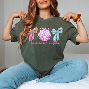 K&ouml;nnte beinhalten: Dunkelgr&uuml;nes T-Shirt mit einer Muschel-Grafik, einer Perle und drei rosa, gelben und blauen Schleifen. Der Text "summer vibes" ist in rosa Schrift geschrieben. Das Shirt besteht aus einem weichen Material. Ein l&auml;ssiges Sommertop.