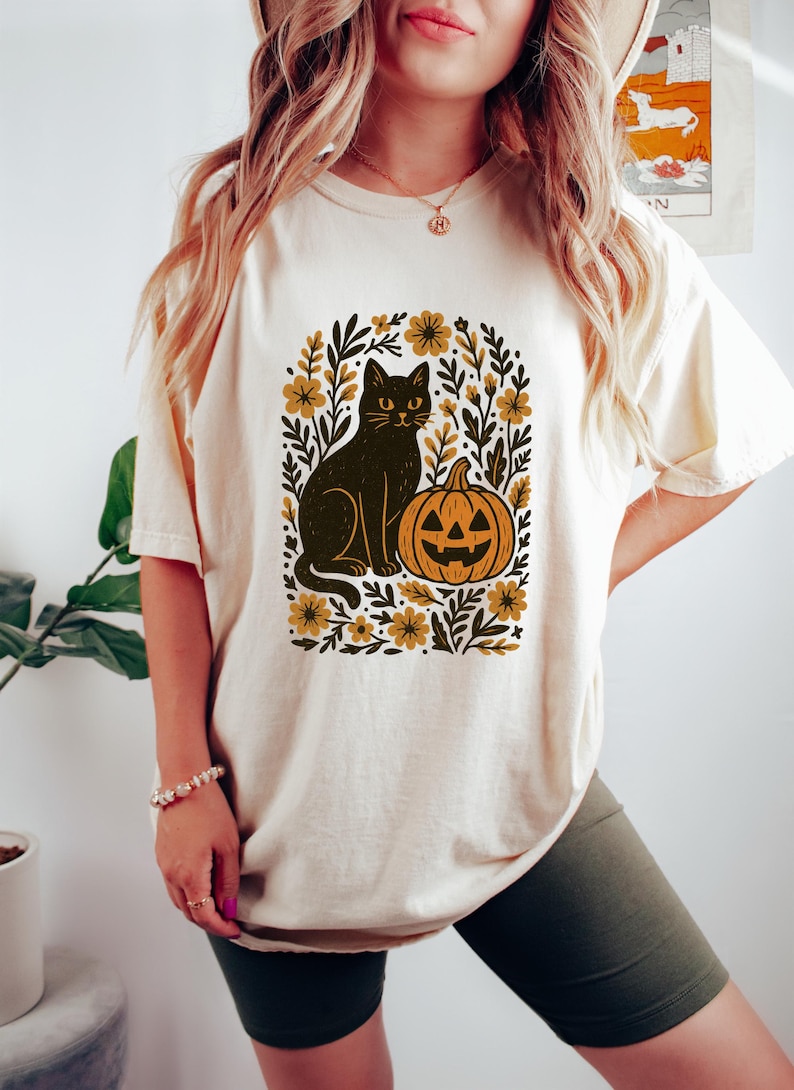 May include: Cream-colored t-shirt featuring a black cat and jack-o'-lantern design. The graphic is surrounded by floral and leaf elements in shades of gold and brown. A Halloween-themed tee.